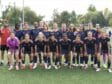 Girls' soccer team posing for a group photo on a green field in navy blue and orange uniforms.