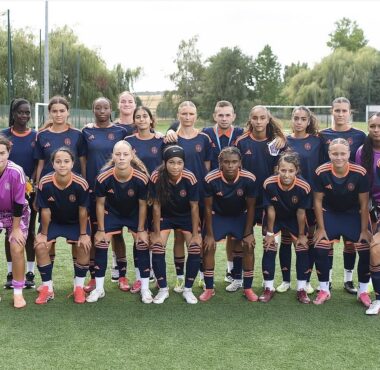Girls' soccer team posing for a group photo on a green field in navy blue and orange uniforms.
