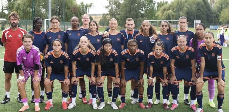 Girls' soccer team posing for a group photo on a green field in navy blue and orange uniforms.