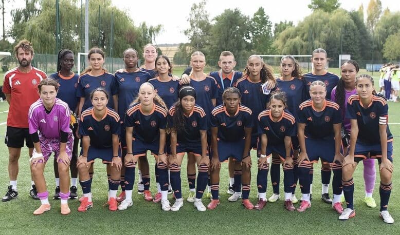 Girls' soccer team posing for a group photo on a green field in navy blue and orange uniforms.