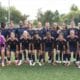 Girls' soccer team posing for a group photo on a green field in navy blue and orange uniforms.