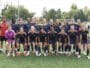 Girls' soccer team posing for a group photo on a green field in navy blue and orange uniforms.
