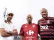 Three men pose for a photo outdoors, wearing soccer jerseys; left in a white polo and cap, center in a red Carrefour City jersey, right in a red-and-black Flamengo jersey with a stadium light in the background.