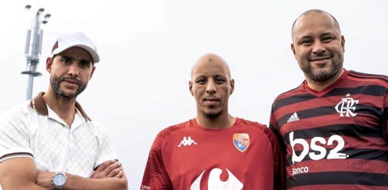 Three men pose for a photo outdoors, wearing soccer jerseys; left in a white polo and cap, center in a red Carrefour City jersey, right in a red-and-black Flamengo jersey with a stadium light in the background.