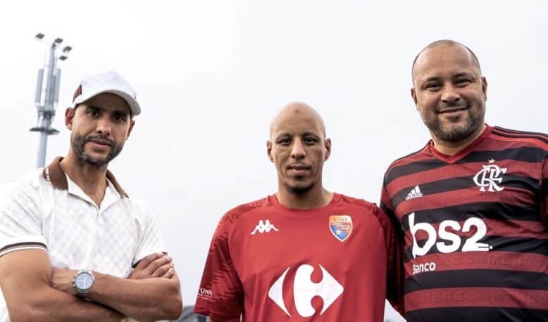 Three men pose for a photo outdoors, wearing soccer jerseys; left in a white polo and cap, center in a red Carrefour City jersey, right in a red-and-black Flamengo jersey with a stadium light in the background.