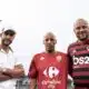 Three men pose for a photo outdoors, wearing soccer jerseys; left in a white polo and cap, center in a red Carrefour City jersey, right in a red-and-black Flamengo jersey with a stadium light in the background.