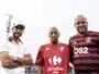 Three men pose for a photo outdoors, wearing soccer jerseys; left in a white polo and cap, center in a red Carrefour City jersey, right in a red-and-black Flamengo jersey with a stadium light in the background.