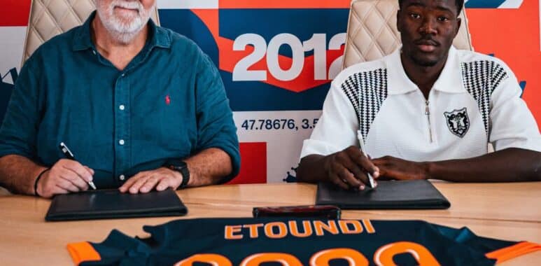 Two men sign documents at a table in front of a colorful sports-themed mural.