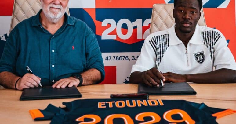 Two men sign documents at a table in front of a colorful sports-themed mural.