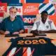 Two men sign documents at a table in front of a colorful sports-themed mural.