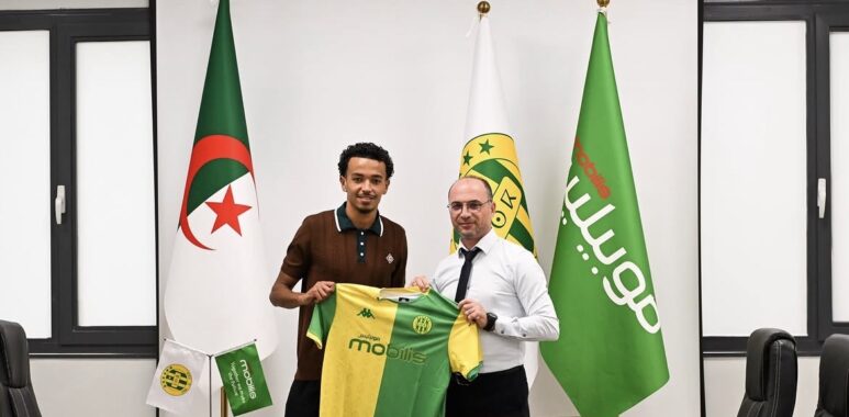 Two men in a formal room hold up a green and yellow football jersey with flags in the background, including the Algerian flag in front of them.