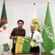 Two men in a formal room hold up a green and yellow football jersey with flags in the background, including the Algerian flag in front of them.