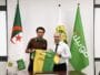Two men in a formal room hold up a green and yellow football jersey with flags in the background, including the Algerian flag in front of them.