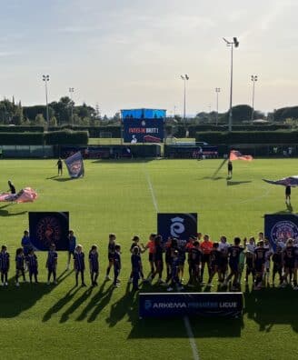 Group of young soccer players in blue uniforms lined up on a sunny green pitch with banners in the foreground and a large scoreboard behind them.
