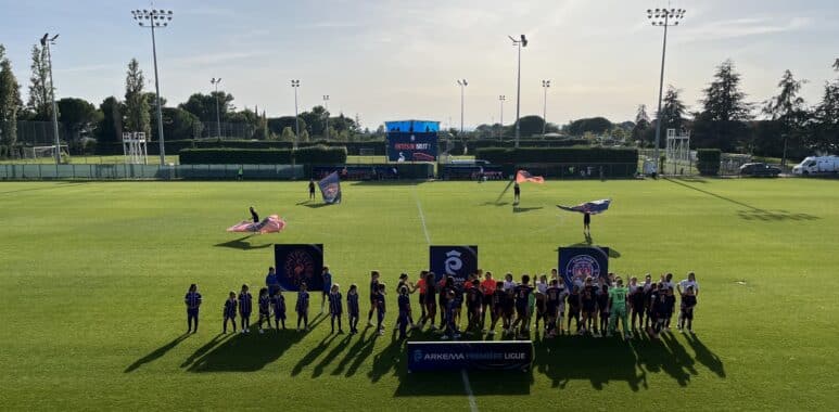 Group of young soccer players in blue uniforms lined up on a sunny green pitch with banners in the foreground and a large scoreboard behind them.