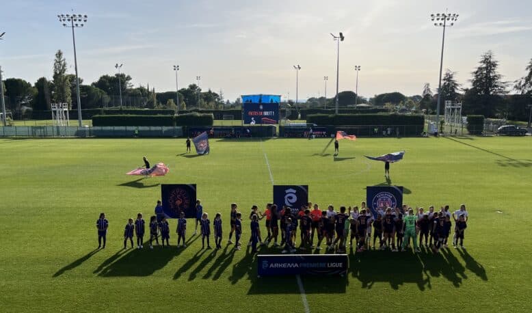 Group of young soccer players in blue uniforms lined up on a sunny green pitch with banners in the foreground and a large scoreboard behind them.