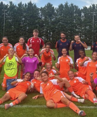 Youth soccer team posing with fists up after a game on a grassy field, orange uniforms with a few in neon vests.