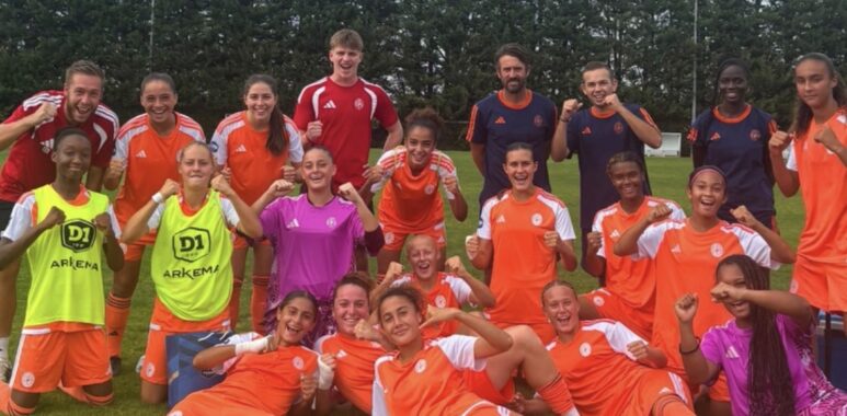 Youth soccer team posing with fists up after a game on a grassy field, orange uniforms with a few in neon vests.