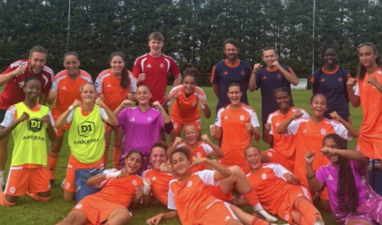 Youth soccer team posing with fists up after a game on a grassy field, orange uniforms with a few in neon vests.