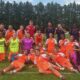 Youth soccer team posing with fists up after a game on a grassy field, orange uniforms with a few in neon vests.
