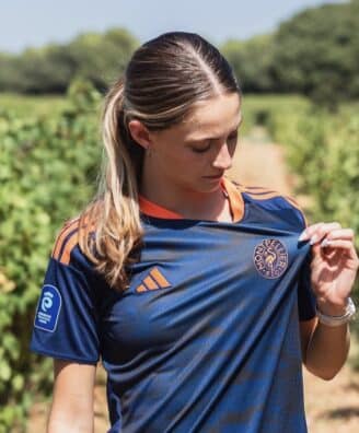Woman in a navy blue sports jersey with orange accents stands in a vineyard, looking down as she adjusts her shirt.