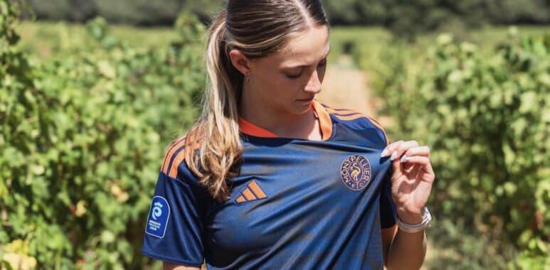 Woman in a navy blue sports jersey with orange accents stands in a vineyard, looking down as she adjusts her shirt.