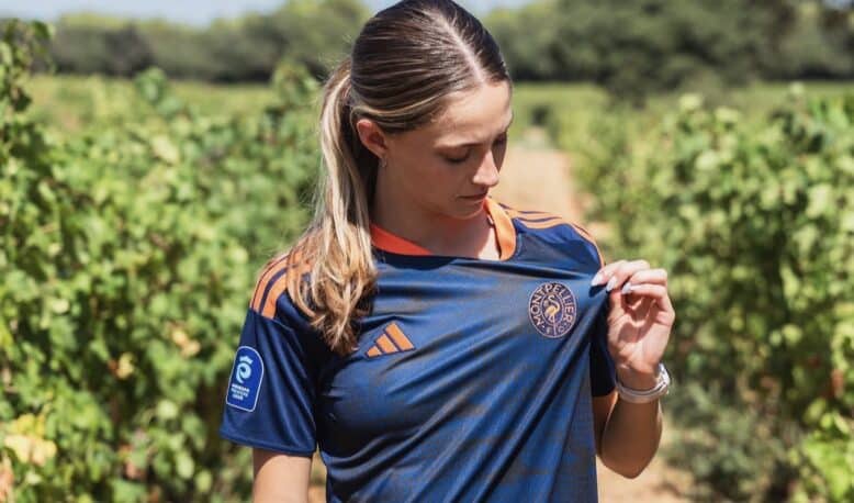 Woman in a navy blue sports jersey with orange accents stands in a vineyard, looking down as she adjusts her shirt.