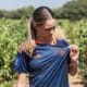 Woman in a navy blue sports jersey with orange accents stands in a vineyard, looking down as she adjusts her shirt.