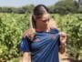 Woman in a navy blue sports jersey with orange accents stands in a vineyard, looking down as she adjusts her shirt.