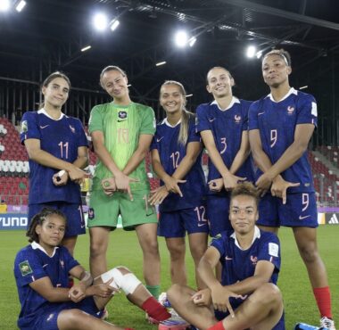 Group of female soccer players posing for a team photo on the field, wearing blue uniforms with numbers and one green goalkeeper in center.