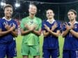 Four female French soccer players stand on a stadium pitch, smiling and crossing their arms over their chests in a show of unity.