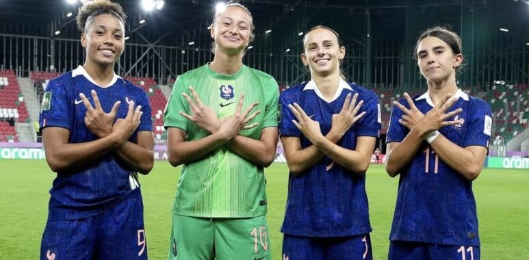 Four female French soccer players stand on a stadium pitch, smiling and crossing their arms over their chests in a show of unity.