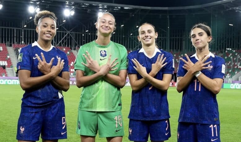 Four female French soccer players stand on a stadium pitch, smiling and crossing their arms over their chests in a show of unity.