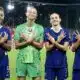 Four female French soccer players stand on a stadium pitch, smiling and crossing their arms over their chests in a show of unity.