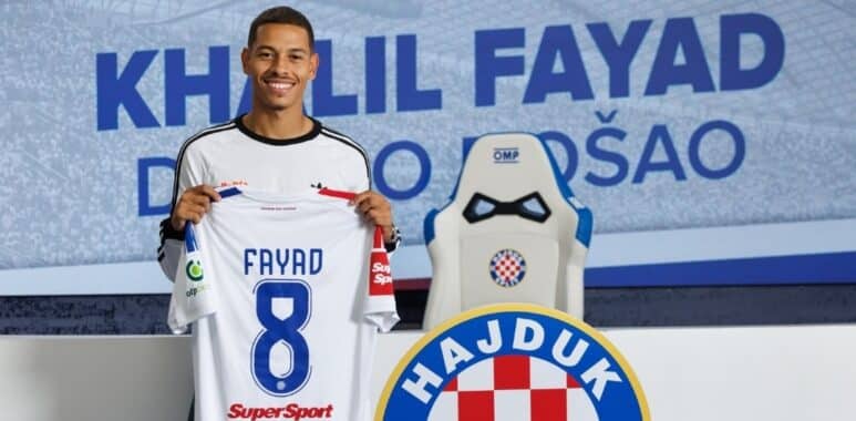 Smiling soccer player holds up a white jersey reading FAYAD 8, in a club press area with Hajduk crest in the background.