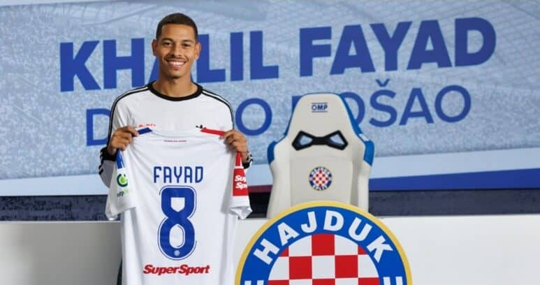 Smiling soccer player holds up a white jersey reading FAYAD 8, in a club press area with Hajduk crest in the background.