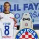 Smiling soccer player holds up a white jersey reading FAYAD 8, in a club press area with Hajduk crest in the background.