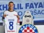 Smiling soccer player holds up a white jersey reading FAYAD 8, in a club press area with Hajduk crest in the background.