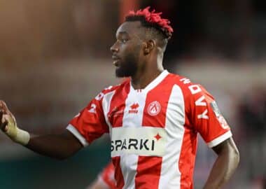 Profile view of a Black male soccer player with a red-tinted mohawk and beard, wearing a red-and-white striped jersey during a match.