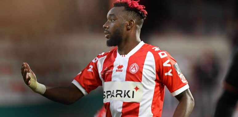 Profile view of a Black male soccer player with a red-tinted mohawk and beard, wearing a red-and-white striped jersey during a match.