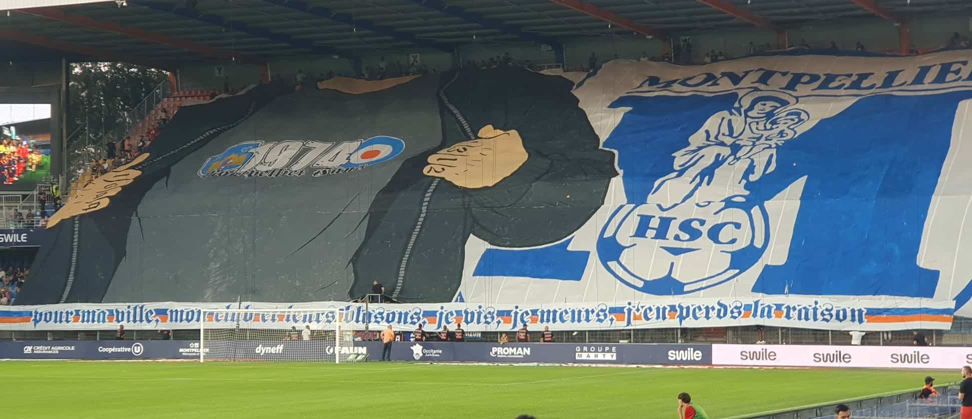 Huge stadium banner showing a grayscale figure with a raised fist and blue graphic elements, behind a football goal and advertising boards.